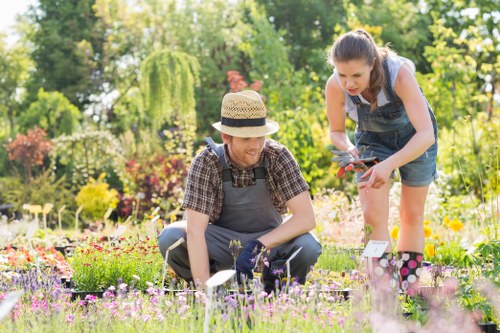 A gardener and a young girl are working together in a vibrant, well-maintained garden with a variety of flowering plants and greenery. The gardener, wearing a straw hat, a checkered shirt, and a grey apron, is kneeling on the lush, densely vegetated lawn, tending to the colorful flowers with gardening tools. The girl, dressed in a denim dress and white t-shirt, is standing nearby, leaning in slightly as she looks at the plants, holding a small garden implement. The garden features a mix of flower beds filled with pink, purple, and white blossoms arranged along the edges of grassy areas, with dense trees and shrubbery providing a lush backdrop. Soft natural sunlight illuminates the scene, highlighting the vibrant colours and textures of the flowers, grass, and foliage. The environment appears to be a peaceful, outdoor residential garden, typical of Hackney or nearby London suburbs, dedicated to outdoor gardening and plant care, with the participation of Landscapers Hackney providing a lush, lively outdoor setting suitable for gardening and landscaping services.
