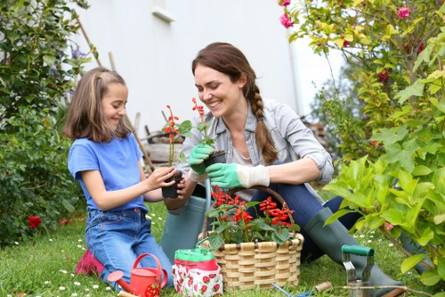 A woman and a young girl are gardening together in a lush backyard garden. The woman, dressed in casual outdoor clothing and green gardening gloves, is smiling as she holds a small potted plant with red flowers, which she appears to be planting or tending to. The girl, wearing a blue t-shirt and denim shorts, is sitting on the grass, also holding the pot and engaging with the gardening activity. Surrounding them are various garden elements, including a wicker basket, a red watering can, a small gardening trowel, and a watering jug. The garden features a well-maintained grassy lawn, flowering plants, shrubs, and a hedge or border along the background, with some taller plants or trees providing a natural backdrop. Natural daylight illuminates the scene, suggesting a pleasant, mild weather day, creating a vibrant and inviting outdoor environment typical of residential gardening spaces in Hackney. The scene exemplifies outdoor garden maintenance, planting, and family engagement in gardening activities, aligning with professional services offered by Landscapers Hackney in local gardening and landscaping projects.
