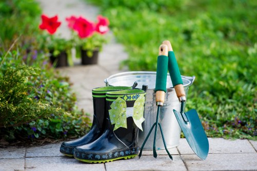 A pair of black rubber gardening boots with green socks draped over them, placed on a paved outdoor surface in a garden setting. Beside the boots, there is a white metal bucket with a pair of garden gloves resting on top, and a small hand trowel with a wooden handle and a blue metal shovel are leaning against the bucket. In the background, vibrant pink and red flowering plants are visible, along with lush green foliage and grass, suggesting a well-maintained garden or backyard. The scene is outdoors on a sunny day, with natural light illuminating the tools and plants, reflecting typical gardening activity in a residential area, possibly in Hackney or nearby in London. The arrangement emphasizes gardening tools and container plants, indicative of ongoing or recent outdoor gardening work, suitable for landscaping or garden maintenance services by Landscapers Hackney.