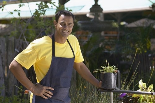 A man wearing a yellow t-shirt and a dark apron stands outdoors in a well-maintained garden, smiling at the camera. Behind him, there are lush green plants, shrubs, and trees, with a garden fountain or sculpture partially visible in the background under a sunny sky. In front of him, on a table, is a small black pot with a young plant, including green leaves and small stems. The garden area features a variety of plantings, natural soil, and paved pathways, creating a neat and inviting outdoor space typical of Hackney gardens, where landscaping and lawn care services by Landscapers Hackney could enhance outdoor environments. The scene suggests a focus on gardening tasks such as planting and garden maintenance, in a setting that combines natural vegetation and landscaped features, appropriate for the local climate and style of the area.