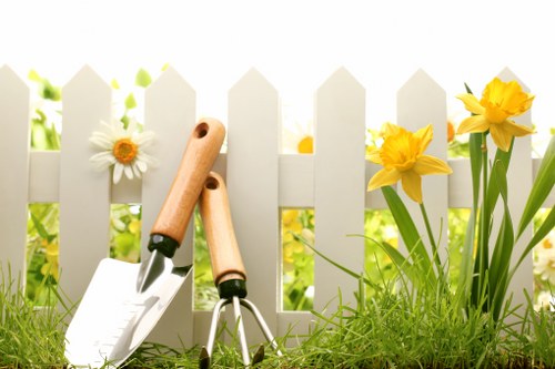 A close-up view of a small suburban garden featuring a white picket fence in the background, with vibrant yellow daffodils and white daisies blooming along the edge. In the foreground, fresh green grass covers the lawn, with two wooden-handled gardening tools—a trowel and a cultivator—resting on the grass near the fence. The garden appears well-maintained, with healthy plants and a neat layout, demonstrating typical features of a landscaped outdoor space in Hackney, suitable for garden care and sustainable planting practices. Natural daylight illuminates the scene, suggesting a clear, mild weather day, emphasizing the importance of outdoor maintenance and sustainable gardening approaches supported by local services such as those offered by Landscapers Hackney.