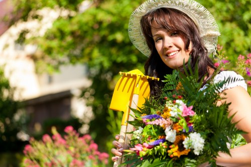 A woman with brunette hair wearing a wide-brimmed straw hat and a white top is standing in a lush garden, holding a vibrant bouquet of mixed flowers and greenery. Behind her, there is a well-maintained lawn with dense, green grass and a pathway or paved area visible at the edge of the garden. In the background, tall hedges and leafy trees frame the scene, with natural sunlight casting a warm glow over the outdoor space. The garden includes flower beds with blooming plants in various shades of pink, purple, white, and yellow, along with neatly edged borders and some shrubbery. The setting suggests a tidy, landscaped garden suitable for outdoor gardening and landscaping services offered by Landscapers Hackney, located in Hackney or nearby London areas. The weather appears pleasant and sunny, enhancing the vibrant colors of the garden. This scene highlights the natural beauty and well-cared-for outdoor environment that outdoor maintenance and planting services can help maintain or create in local gardens.