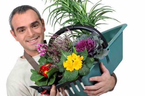 The image shows a smiling man holding a green plastic garden rake filled with a variety of colourful flowering plants, including purple, yellow, and red blooms, set against a bright background. Behind him, a potted plant with long, green, leafy foliage is visible, indicating a well-maintained outdoor garden space. The scene suggests a focus on gardening and outdoor plant care, with the foliage and flowers demonstrating healthy growth typical of landscaped gardens managed by professional landscapers in Hackney. The man's attire and the garden tools hint at professional landscaping or gardening services aimed at enhancing outdoor environments within the Hackney area, including lawn care, planting, and garden maintenance. The natural lighting highlights the vibrant colours and textures of the plants, emphasizing the lush, inviting character of a well-kept outdoor area, closely aligned with local gardening service offerings.