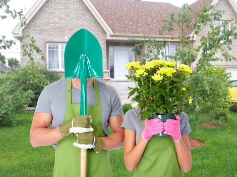 A person and a child, both dressed in grey t-shirts and green gardening aprons, are standing on a well-maintained lawn in front of a suburban house with a pitched roof and brick exterior. The adult is holding a turquoise gardening trowel while wearing beige gardening gloves, and the child is discreetly holding a large potted plant with bright yellow flowers and lush green foliage, partially covering her face. The garden features a neatly trimmed hedge in the background, with the lawn area showing a dense, evenly mown grass surface. There are young trees with light green leaves surrounding the front yard, and the scene is illuminated by natural daylight, suggesting a clear or partly cloudy day. The outdoor space appears tidy and landscaped, reflecting professional gardening practices and sustainable outdoor maintenance supported by Landscapers Hackney.