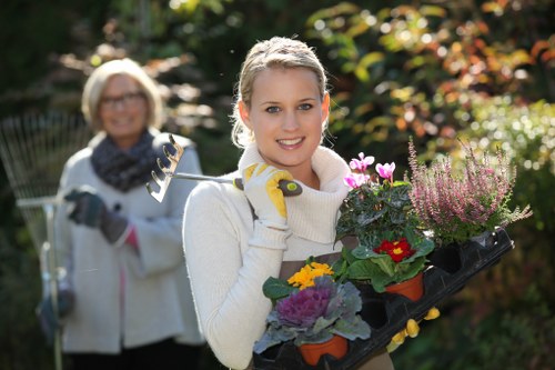 A young woman with blonde hair, wearing a cream-colored sweater and yellow gardening gloves, is holding a black tray filled with colorful potted plants, including purple, yellow, and red flowers, in a well-maintained garden during daylight. Behind her, an older woman with short blonde hair, glasses, and a white coat is holding a garden rake and smiling, standing in front of a lush garden with green foliage, bushes, and trees, which are illuminated by natural sunlight. The garden features a variety of plants, and the scene captures a peaceful outdoor space suited for gardening and landscaping activities near Hackney, with vibrant natural tones and well-organized planting areas.